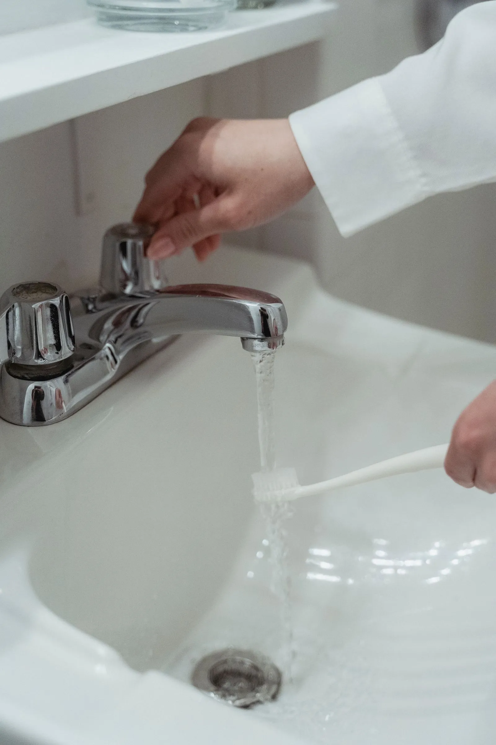 Hand rinsing a white toothbrush under a running tap at a white sink